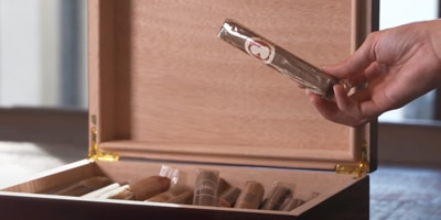 Hand holding a cigar stick wrapped in cellophane above a box of cigars